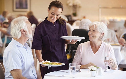 Senior Couple Being Served With Meal By Carer In Dining Room Of Retirement Home